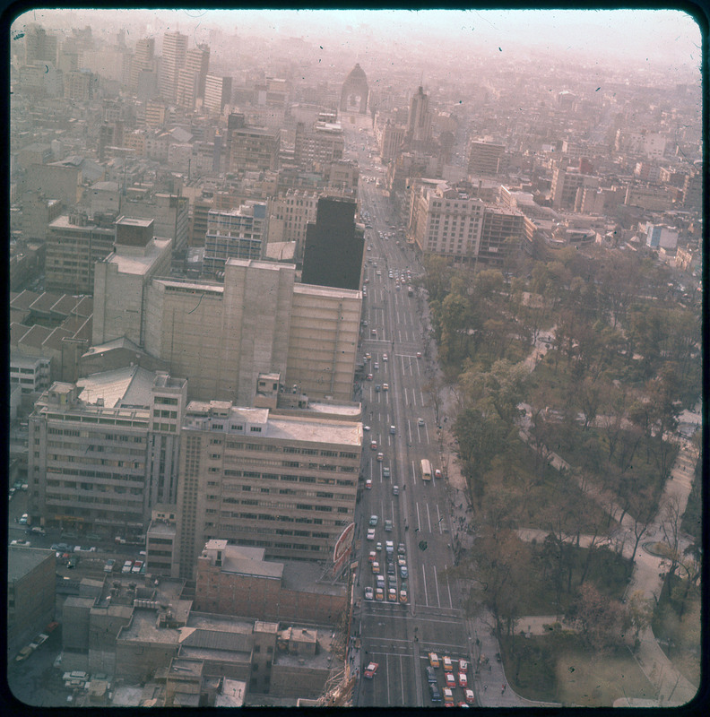 A photographic slide of a large city street of cars. There is a park on one side of the slide and city skyscraper buildings on the other.