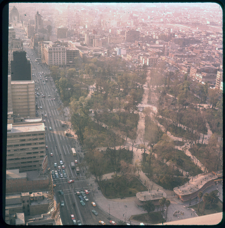 A photographic slide of a large city park with many walkways. There are skyscrapers and a city with roads in the background.