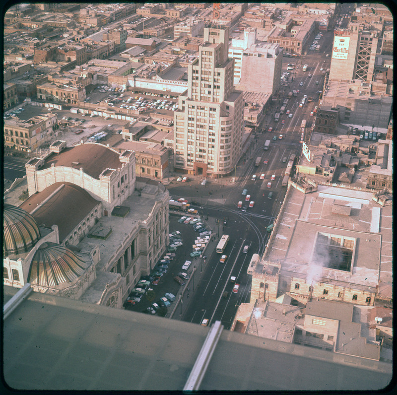 A photographic slide of busy city road and many city buildings. There are cars parked on top of some of the buildings.