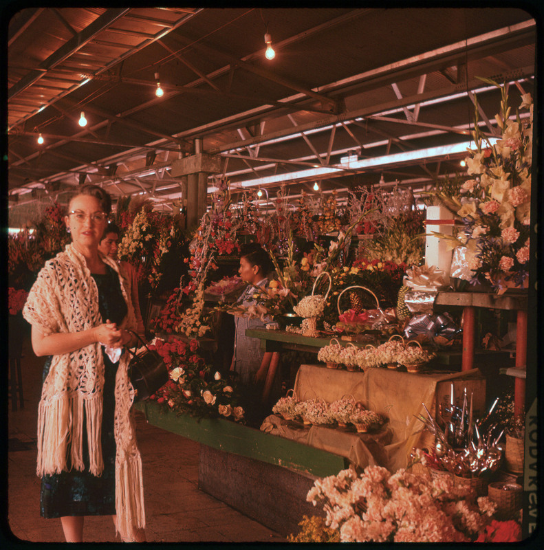 A photographic slide of Evelyn Crabtree standing in front of many flowers at an indoor floral market. The flowers are in arrangements and there is a person selling flowers behind her.