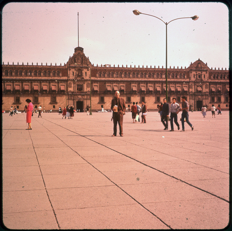 A photographic slide of Donald Crabtree standing in the middle of a courtyard in front of a large building. There are many people in the courtyard.