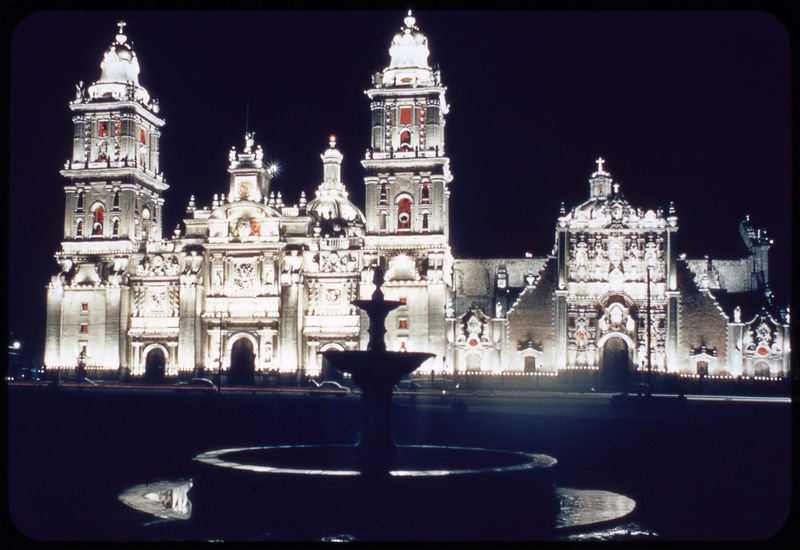 A photographic slide of the Mexico City metropolitan cathedral lit up in lights at night. There is a fountain out front.