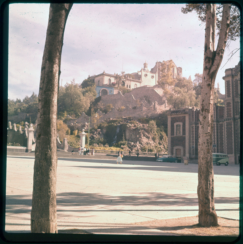 A photographic slide of a large building nestled on a hill with a garden. There is a large staircase out front and people walking in the courtyard.