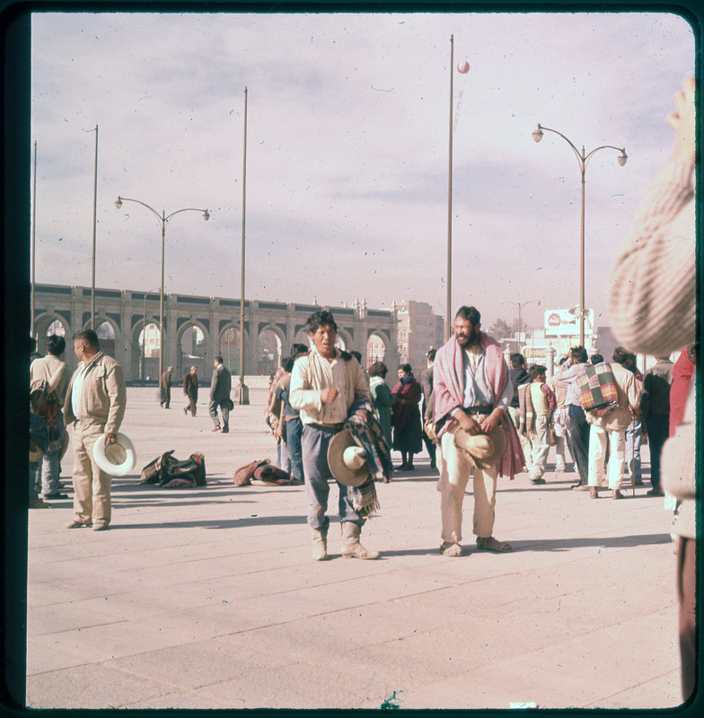 A photographic slide of a courtyard likely in Mexico city. There are many people in the courtyard, and the photo features two people specifically, holding hats and blankets.