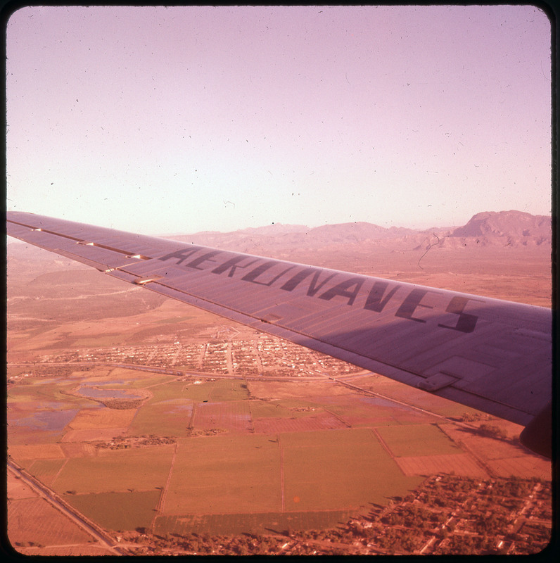 A photographic slide of an airplane wing. In the background there is a city, farmland, and mountains.