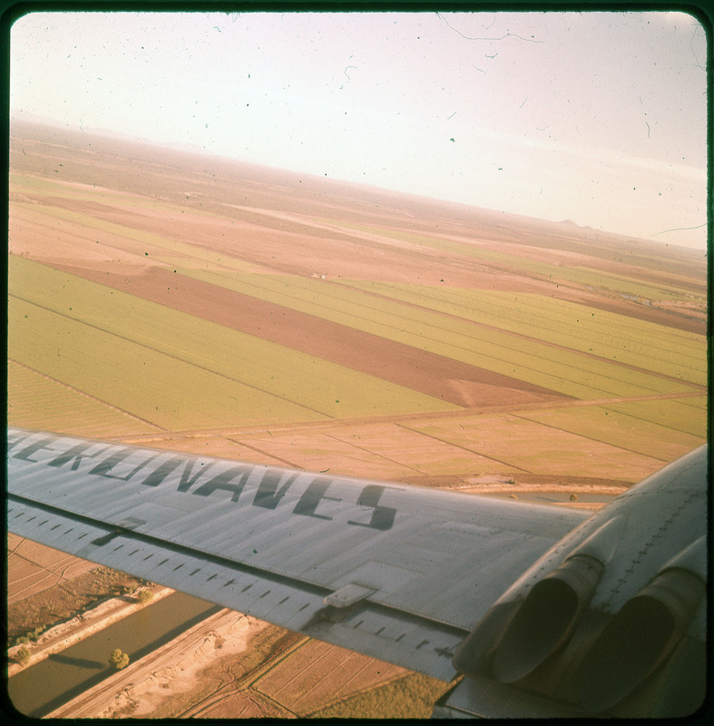 A photographic slide of agricultural fields in slender vertical shape. The wing of an airplane is visible and there are distant mountains in the background.