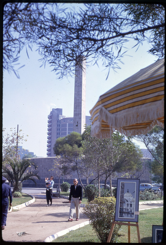 A photographic slide of an outdoor art gallery with a tall, spindly building in the background.