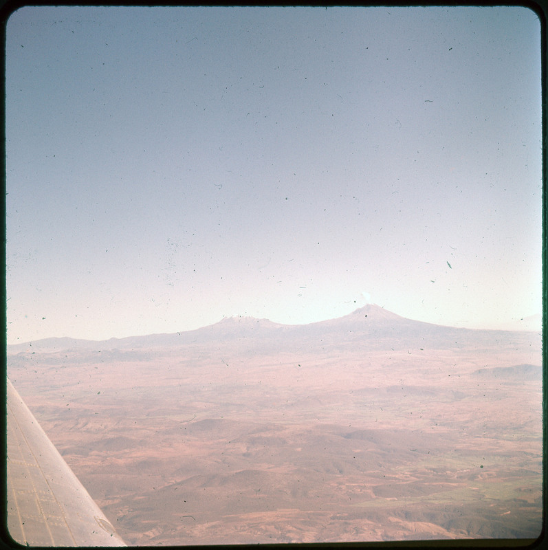 A photographic slide of two volcanoes in the distant, one with smoke coming out. There are hills in the foreground.