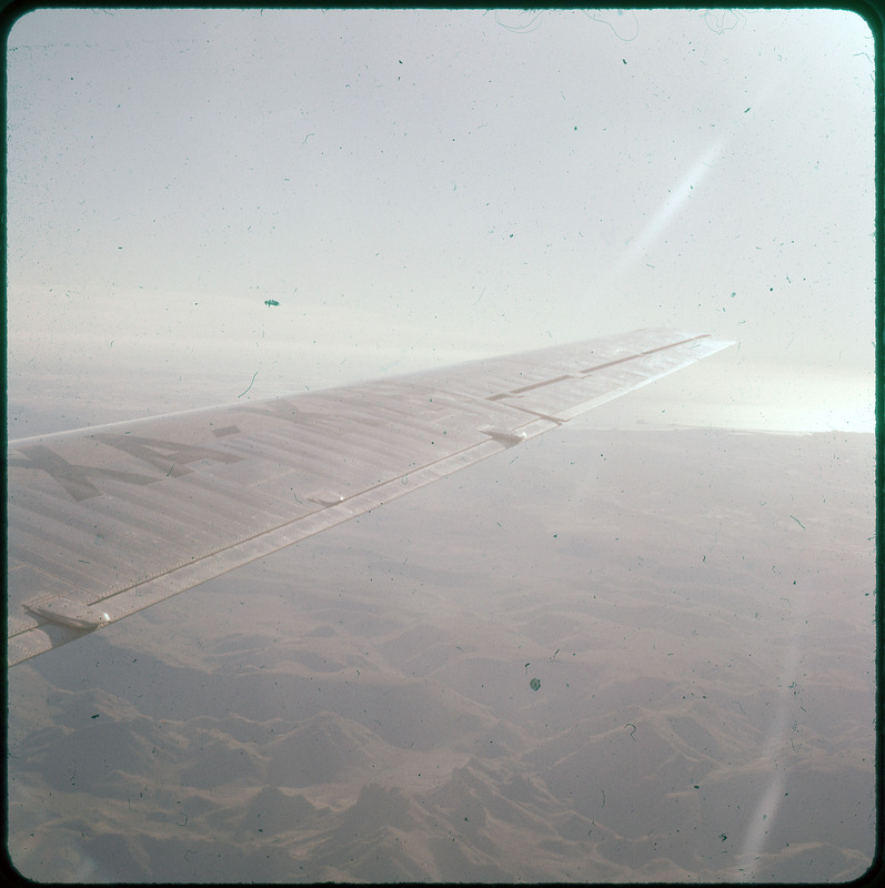 A photographic slide of the wing of an airplane from above. There are mountains in the background and the ocean is visible.