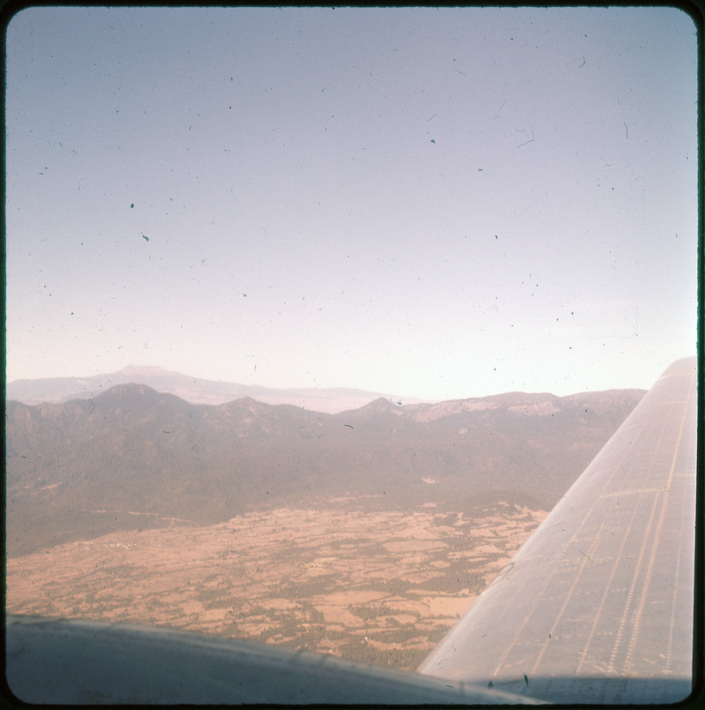 A photographic slide of a a mountain range and field from the window of an airplane in the sky.