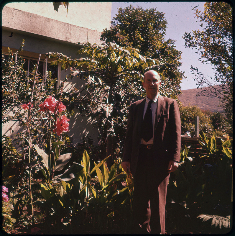 A photographic slide of Donald Crabtree posing in front of plants outdoors. There is a building in the background.