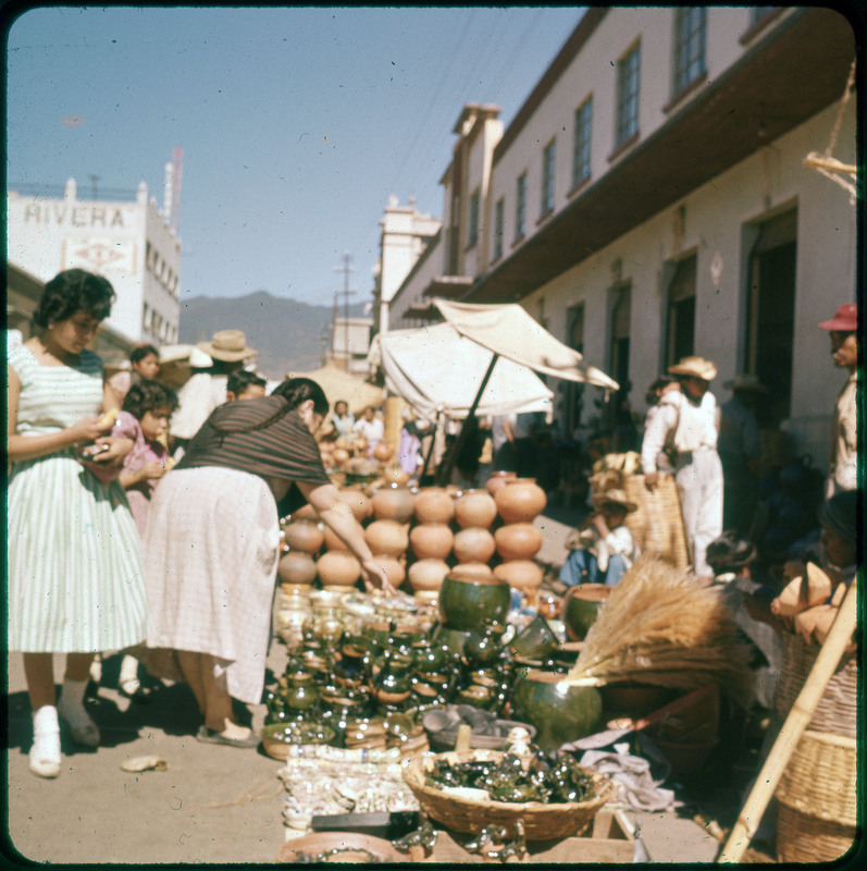 A photographic slide of people shopping in an outdoor market. There are terracotta pots being sold and some other ceramic pots.