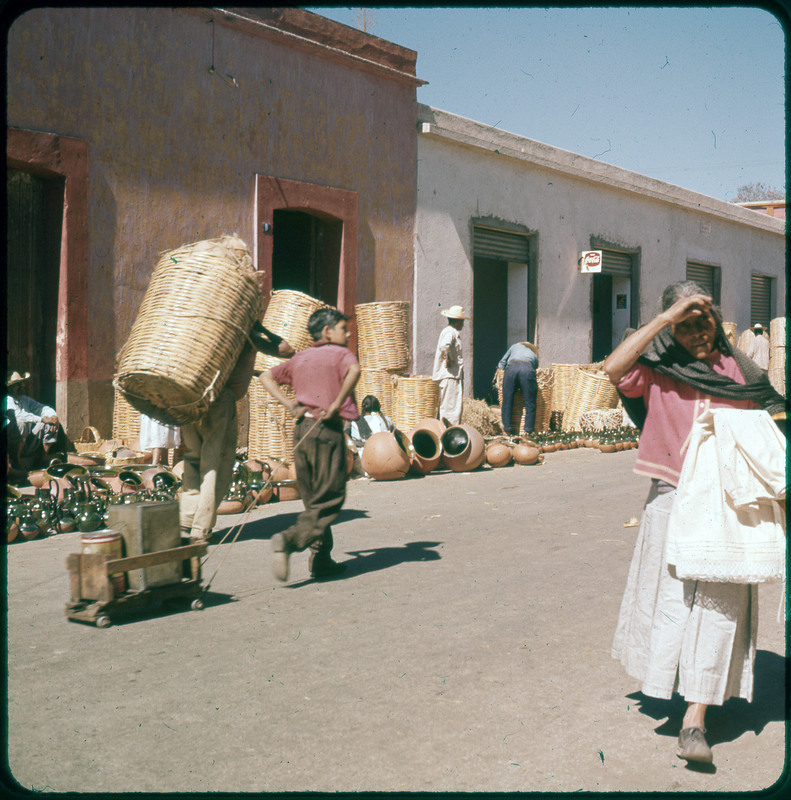 A photographic slide of people shopping in an outdoor market. There are baskets and pots being sold. One person is lugging a cart of materials.