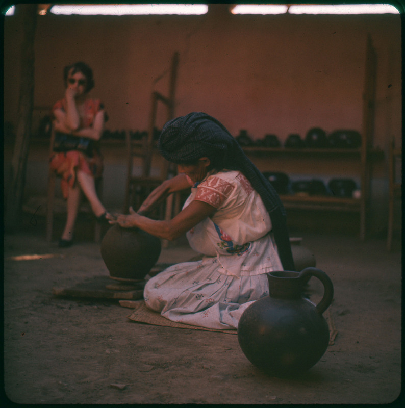 A photographic slide of a person working on making a ceramic pot. There are other pots in the background and a pitcher in the foreground.