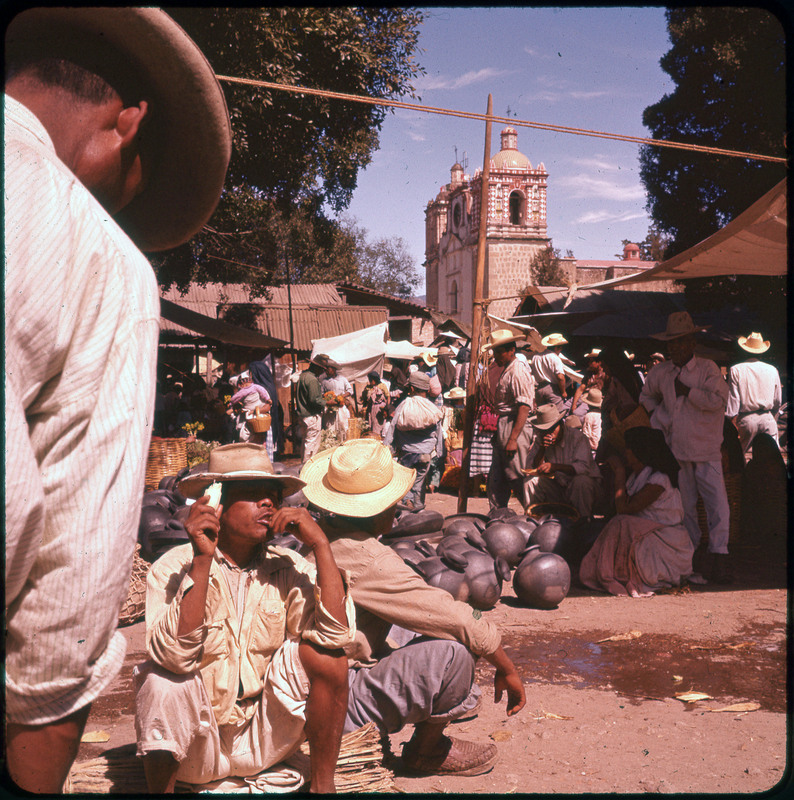 A photographic slide of many people shopping in an outdoor market. There are many pots on the ground and a religious building with crosses in the background.