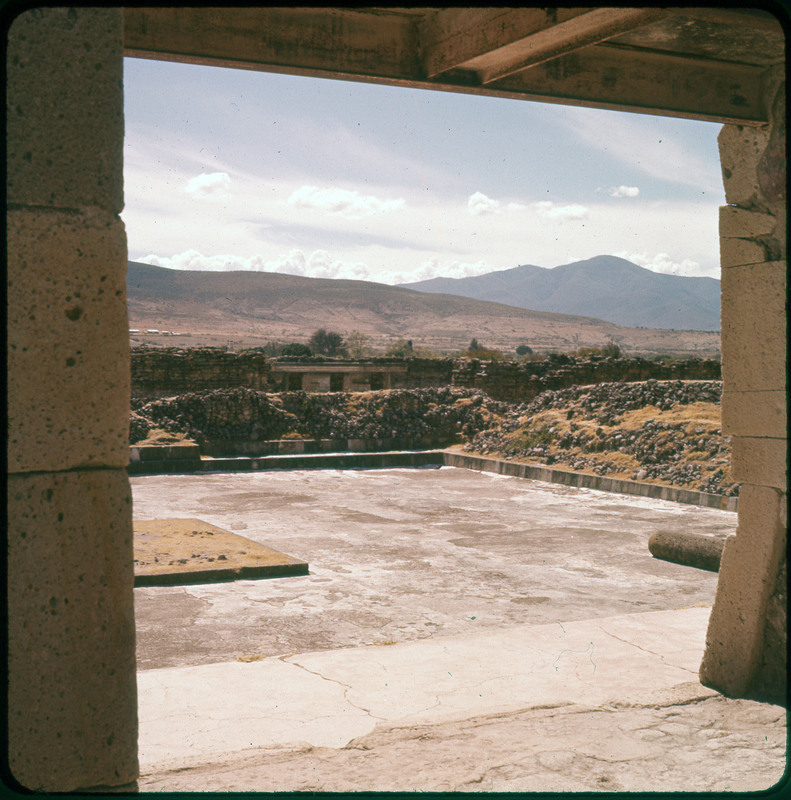 A photographic slide from the view of a stone courtyard with mountains in the background. There are rocky hills in the foreground.