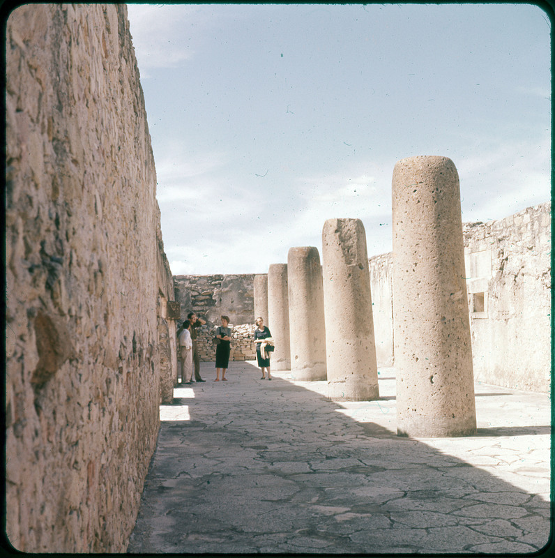 A photographic slide of Evelyn Crabtree and three other people in a stone courtyard. There are five large pillars in and stone walls surrounding them.