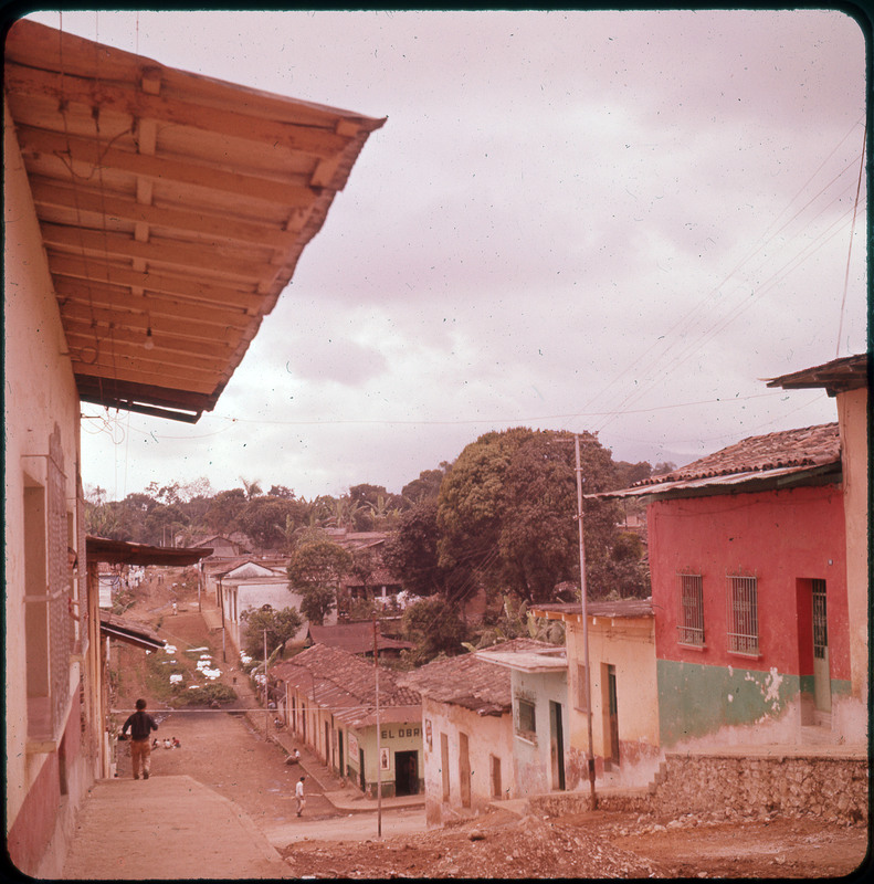 A photographic slide of people walking on a village street of buildings on a hill. There are trees in the background.