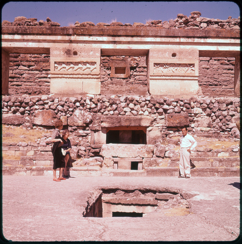 A photographic slide of three people standing in a stone courtyard. There is a large stone wall with unique bricks arranged. There are two holes and the building has an underground.