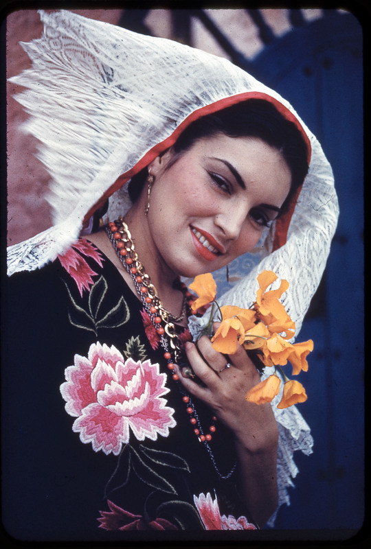 A photographic slide of a woman wearing a headdress, a floral dress, many necklaces and holding yellow flowers.