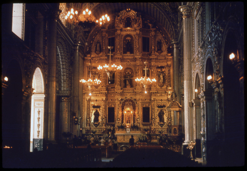 A photographic slide of a golden ornate interior to a church. There are pews, and many ornate decorations. There are people at the front of the room.