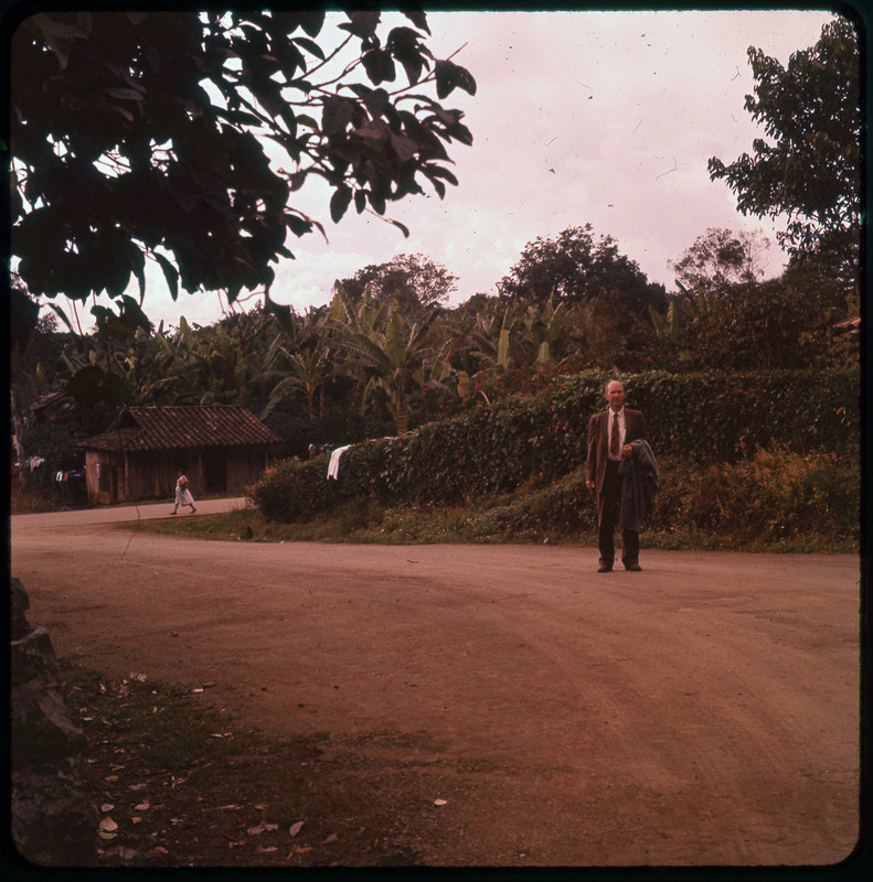 A photographic slide of Donald Crabtree standing on a road in a tropical place. There are hedges in along the road and palm trees in the background. There is a building in the background.