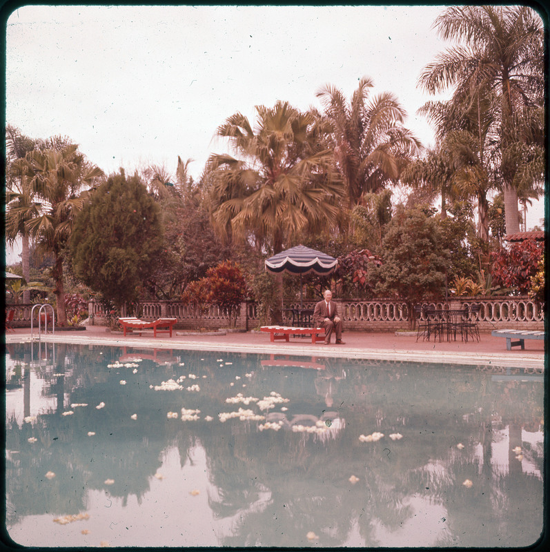 A photographic slide of Donald Crabtree sitting on a bench next to a pool with white flowers in it. There are trees in the background.