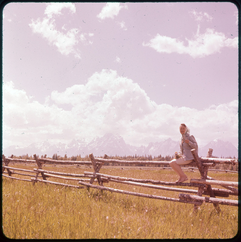 A photographic slide of Evelyn Crabtree sitting on a fence in front of a large field, pine trees, and snow capped mountains in the background.