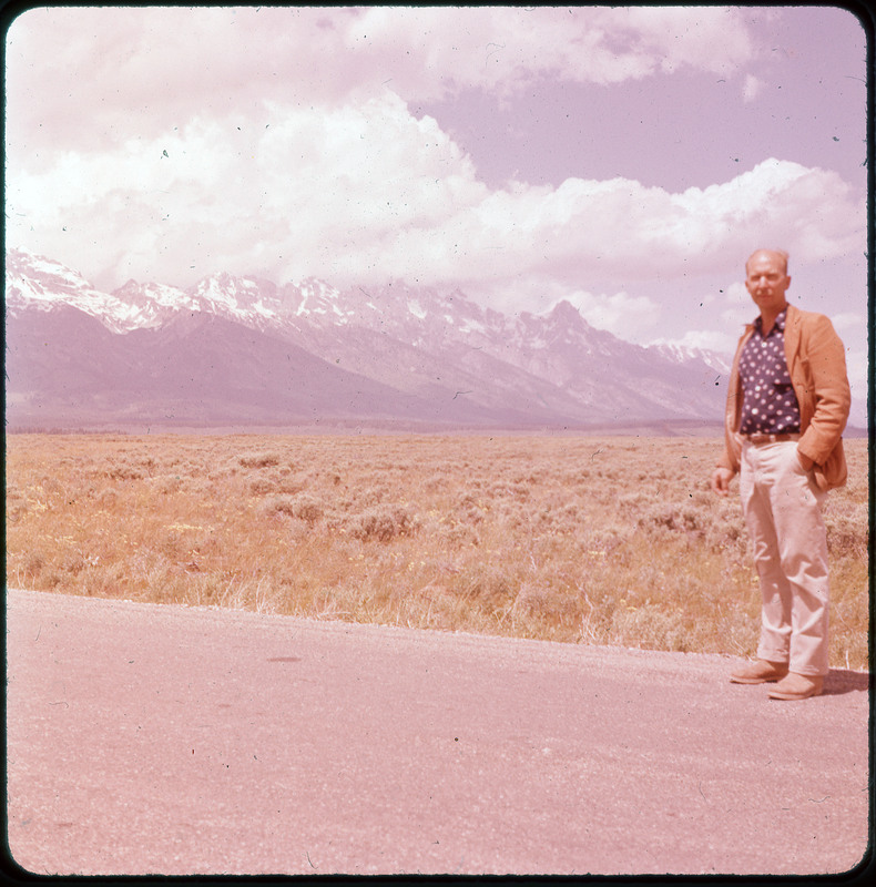 A photographic slide of Donald Crabtree standing in front of a large grassy field. There are snowy mountains in the background.