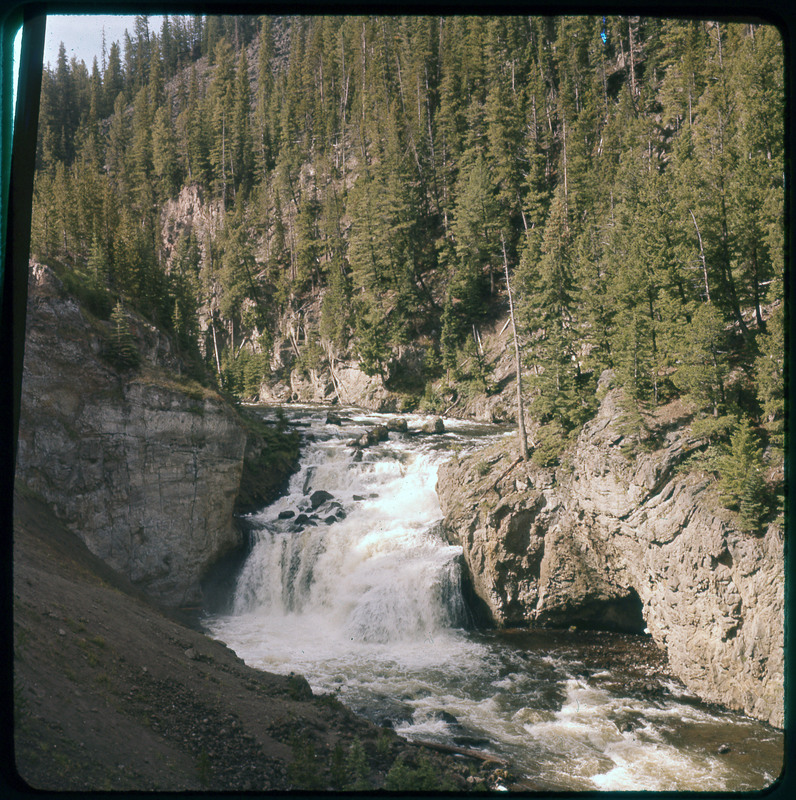 A photographic slide of a river corridor. There is a large waterfall and rocky surroundings. There are pine trees lining the river.