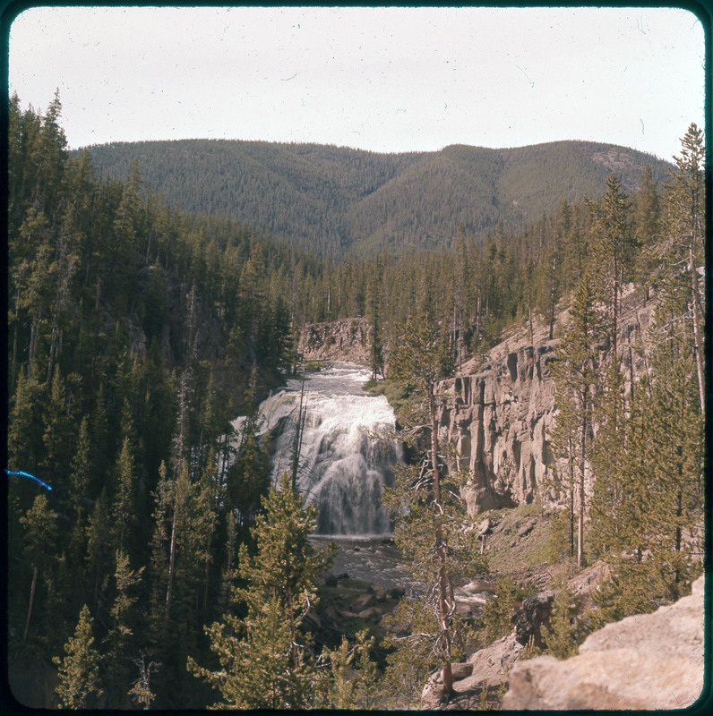 A photographic slide of a river corridor and mountains in the background. There is a large waterfall at the center of the photo and rock faces on the side.