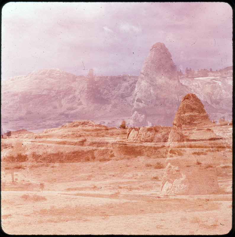 A photographic slide of rock structures, a road, and grassy fields. The slide seems to be two faded photos overlapped. There are faded mountains in the background.