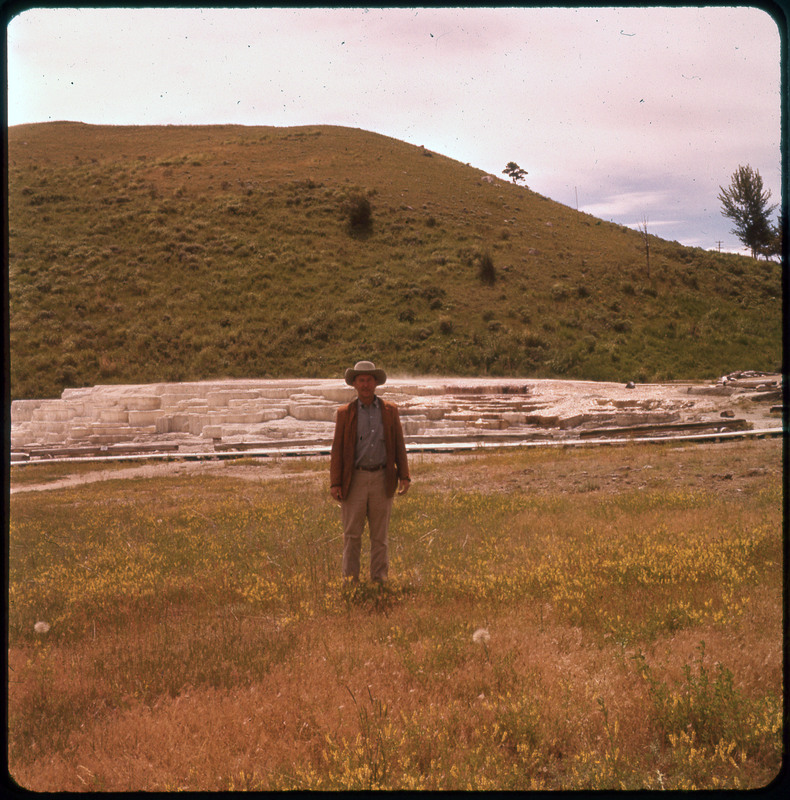 A photographic slide of Donald Crabtree posing in front of a hot pool with brick like rocks stacked. There is a mountain in the background.