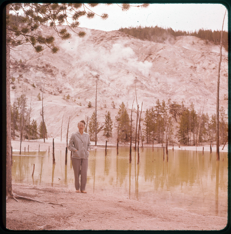 A photographic slide of Evelyn Crabtree posing in front of a green tinted pond with dead trees in it. There is a large snowy mountain in the background.