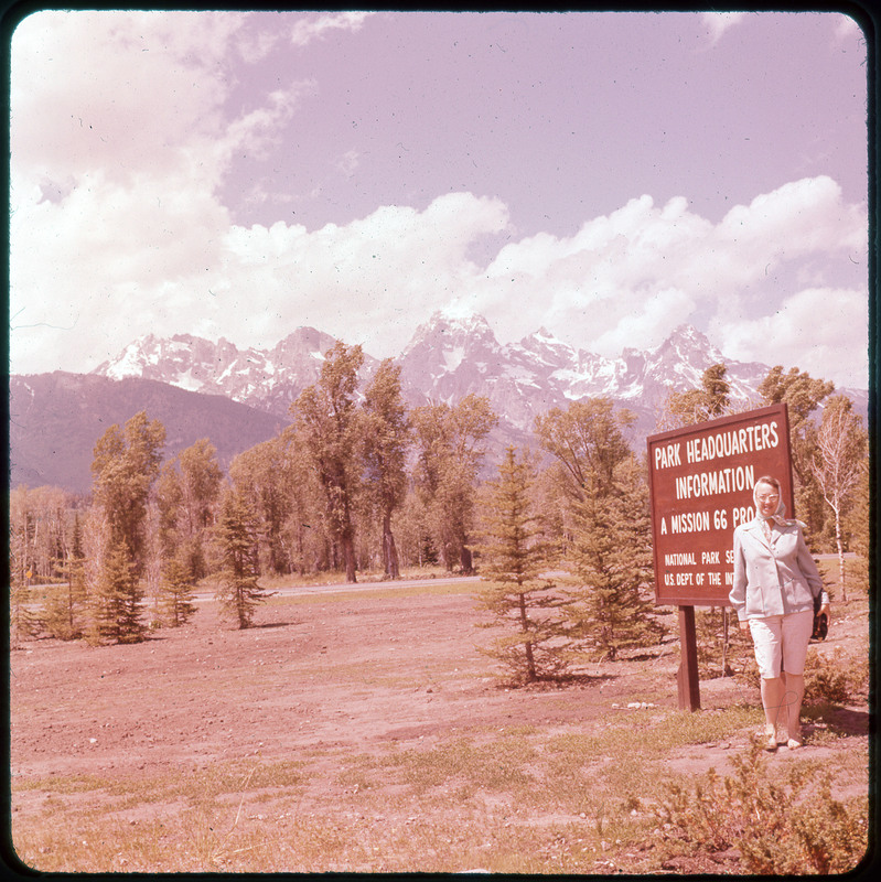 A photographic slide of Evelyn Crabtree next to a national park headquarters sign. There are snowy mountains in the background, a road and many pine trees.