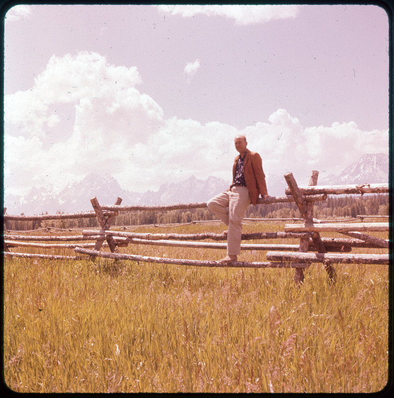 A photographic slide of Donald Crabtree sitting on a fence in front of a grassy field. There are snowy mountains in the background and pine trees.