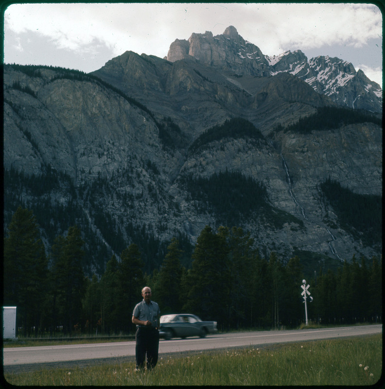 A photographic slide of Donald Crabtree standing in front of a rocky large mountain. There are pine trees in the foreground.