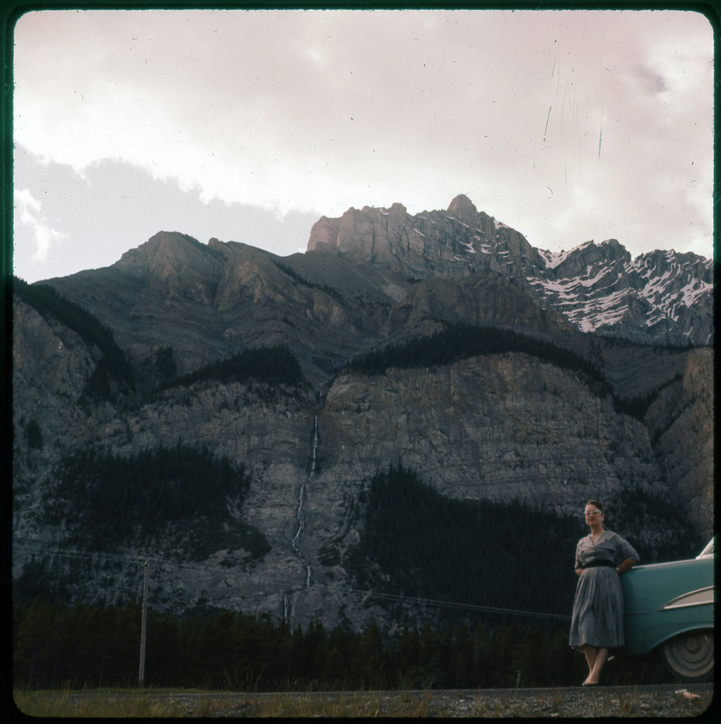 A photographic slide of Evelyn Crabtree standing in front of large rock faces and mountains. There are pine trees on parts of the rock and a waterfall.