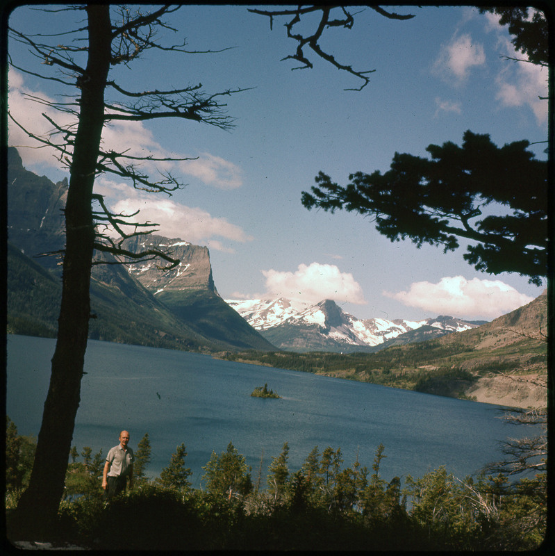 A photographic slide of Donald Crabtree standing in front of a large lake. There are many snowy mountains in the background and pine trees.