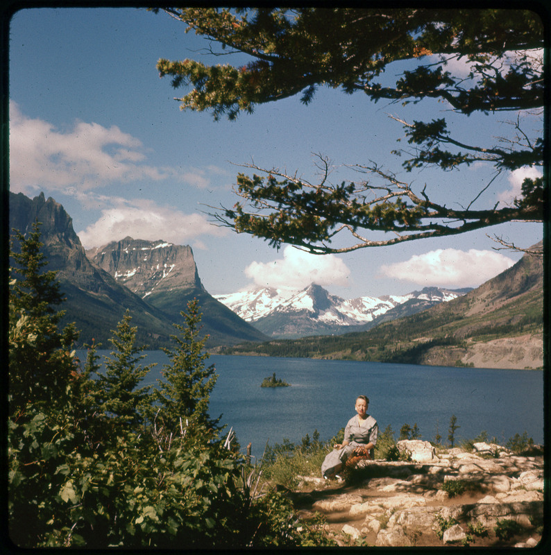 A photographic slide of Evelyn Crabtree standing in front of a large lake. There are many snowy mountains in the background and pine trees.