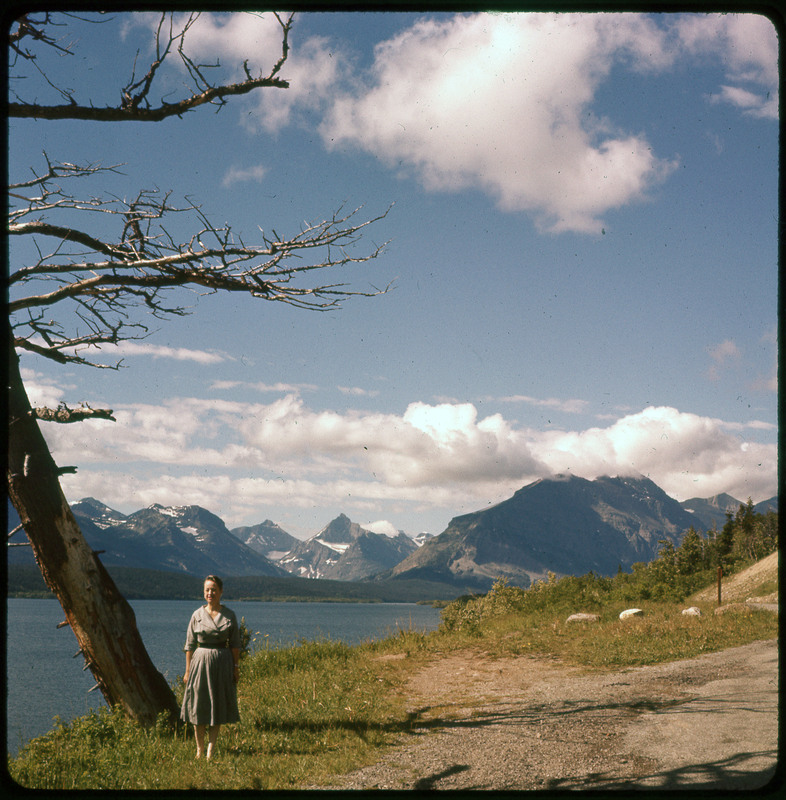 A photographic slide of Evelyn Crabtree standing next to a tree. There is a large lake and snowy mountains in the background.