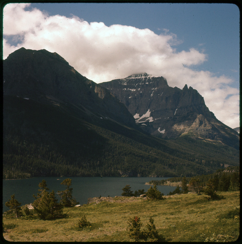 A photographic slide of a large lake in front of snowy mountains. There are many pine trees.