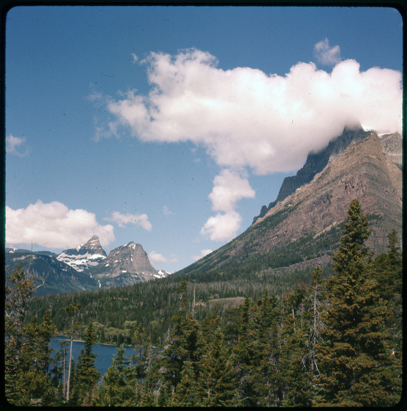 A photographic slide of mountains with many pine trees and a clouds in the sky.