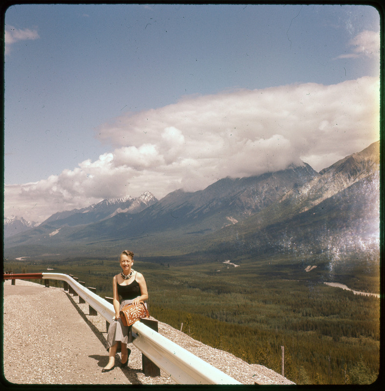 A photographic slide of Evelyn Crabtree sitting on a guardrail in front of a large snowy mountain range. There is a forest in the background.