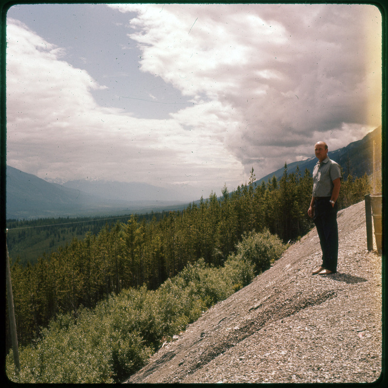 A photographic slide of Donald Crabtree on a hillside overlooking a pine tree forest with many snowy mountains in the background.