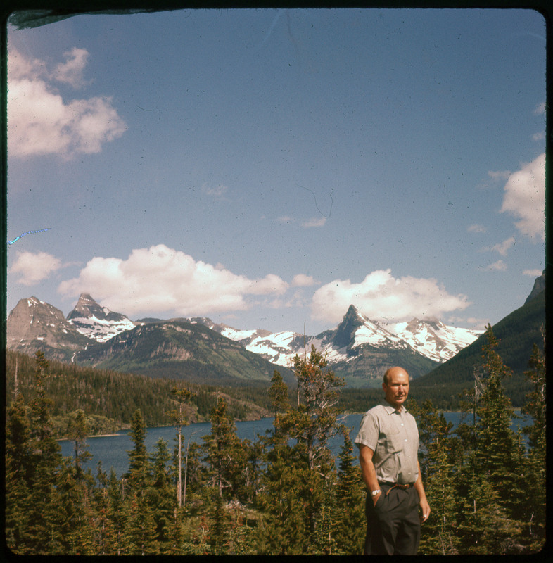 A photographic slide of a snowy mountain range and cloudy sky. Donald Crabtree is posing in front and there is a lake visible.