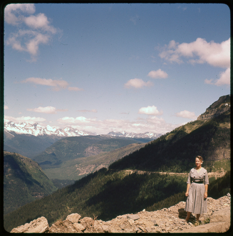 A photographic slide of a large forested valley with Evelyn Crabtree posing in front. There is a snowy mountain range in the background.