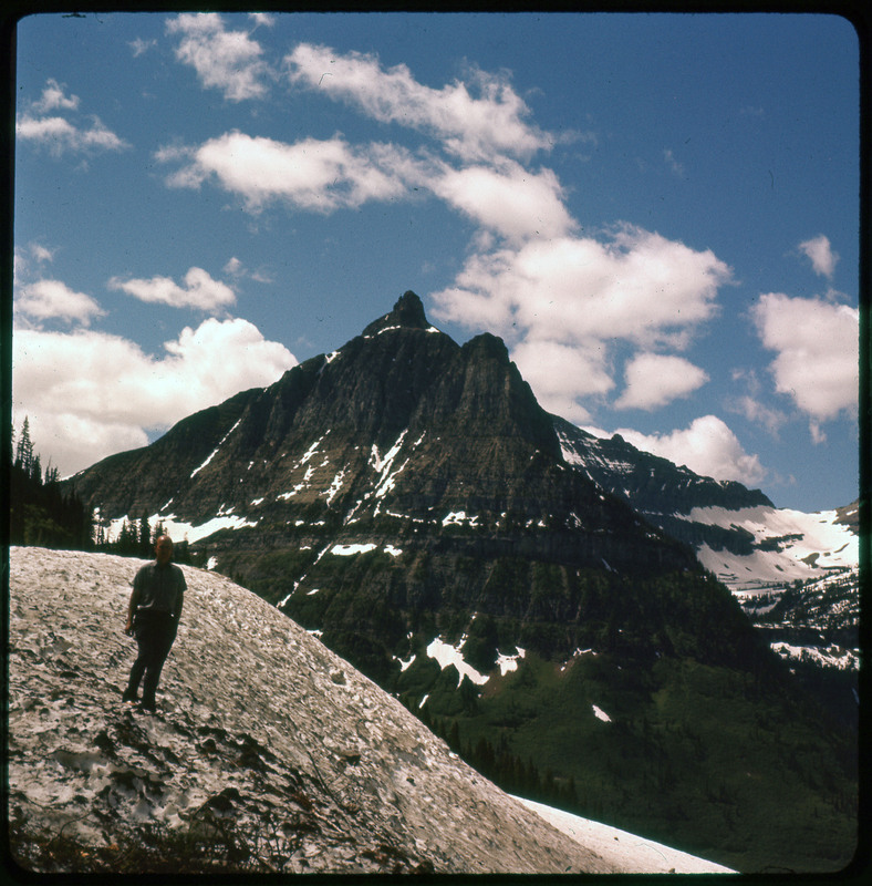 A photographic slide of Donald Crabtree on a rock standing in front of a large mountain range. The mountains have some snow on them.