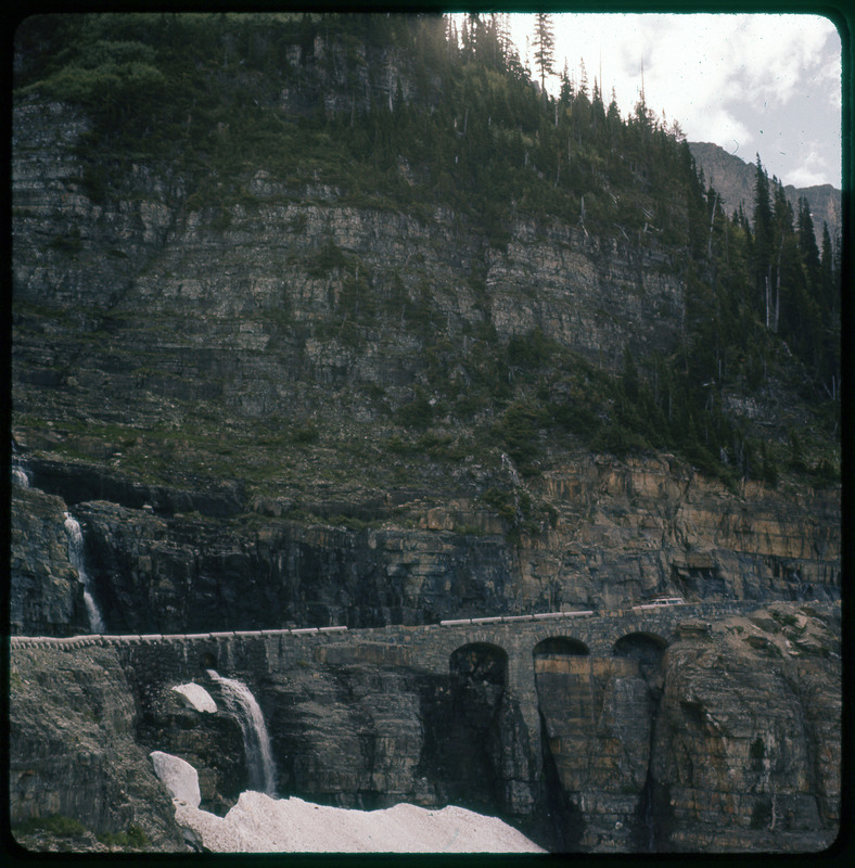 A photographic slide of a car on a mountain road. There is a large rocky mountain pictured with a small waterfall.