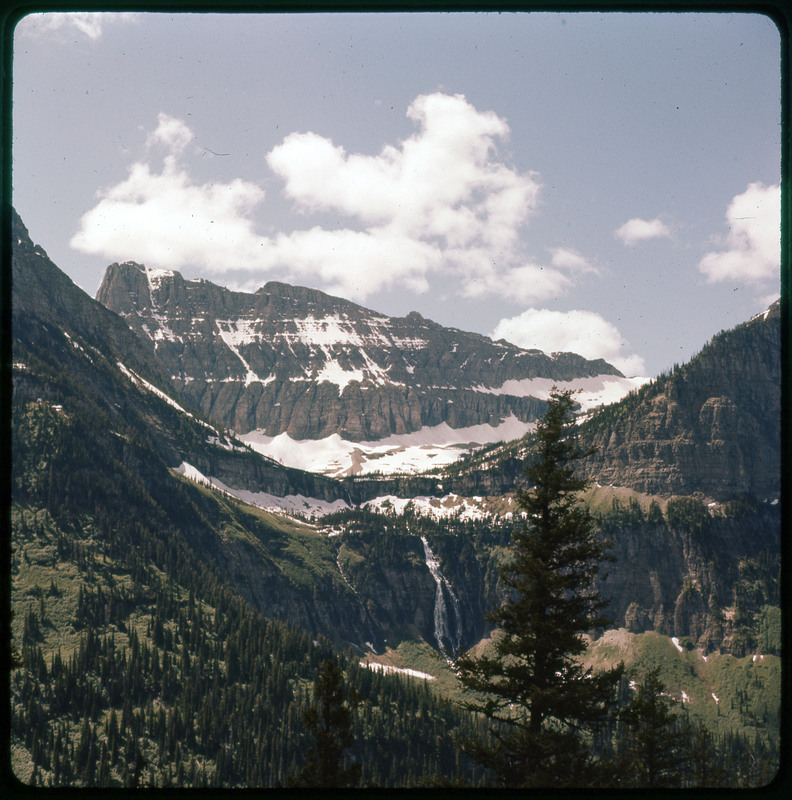 A photographic slide of a waterfall in front of a large snowy mountain range and valley.